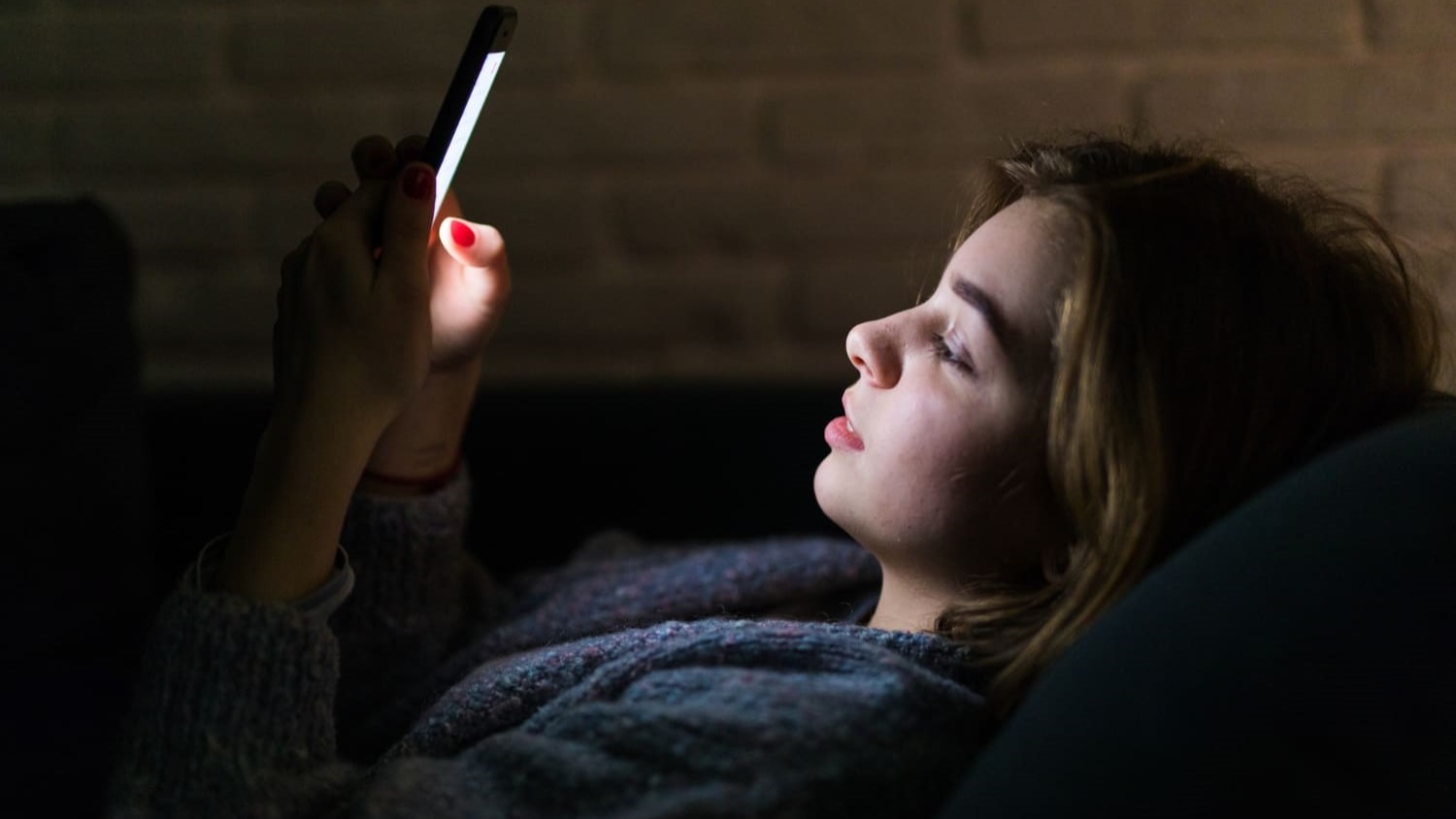 Meisje ligt in bed en kijkt in het donker naar haar telefoon, wat een ongezonde schermroutine benadrukt.
