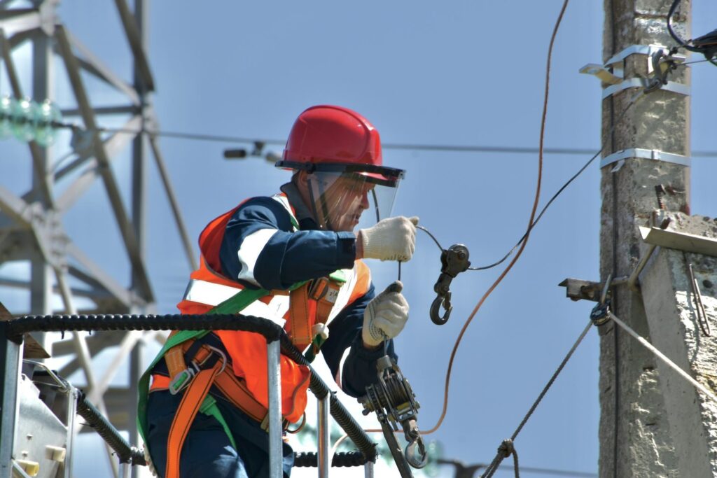 Monteur met helm en veiligheidsharnas inspecteert elektriciteitskabel op hoogte om kortsluiting te voorkomen.
