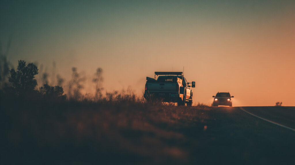 Een pickup met dakverlichting staat stil langs de weg bij zonsondergang terwijl een tegemoetkomende auto met koplampen nadert op de landweg